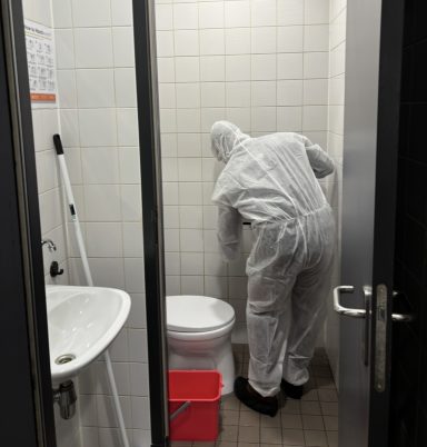 Een persoon in een beschermend pak reinigt een badkamer met een toilet en wastafel.