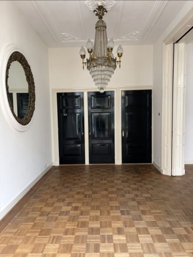 Licht interieur met houten vloer, spiegel en een kristallen kroonluchter aan het plafond.