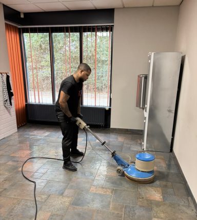 Een man schoonmaakt een vloertegel met een schrobmachine in een lege ruimte.